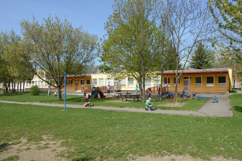Školka Sluníčko – pohled na zahradu mateřské školy s klouzačkou, pískovištěm  a zpevněným povrchem pro odrážedla v pozadí budova mateřské školy Školní 1804  